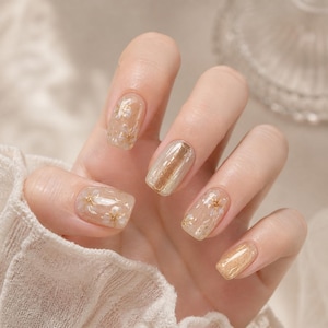 Puede incluir: Una mano con uñas cuidadas. Las uñas presentan una variedad de diseños, incluyendo esmalte transparente con detalles dorados, purpurina dorada y una raya dorada. Algunas uñas tienen pequeños detalles de flores blancas. La estética general es elegante y femenina.