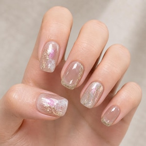 Puede incluir: Una mano que muestra una manicura con esmalte de uñas transparente y color nude. Algunas uñas presentan detalles de purpurina dorada y diseños florales rosas. Las uñas son una mezcla de color sólido y diseños decorados, mostrando un estilo delicado y elegante.