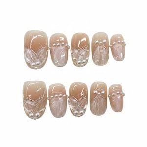 Puede incluir: Un juego de diez uñas artificiales de varios tamaños. Cada uña presenta una base nude con un diseño de remolino nacarado y un acento blanco en forma de hoja. Pequeñas perlas blancas están dispuestas en la base de cada uña.