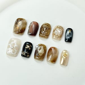 May include: Set of ten false nails with various designs. The nails are decorated with gold, black, white, and brown accents. Some nails feature swirls, seashells, and floral patterns.