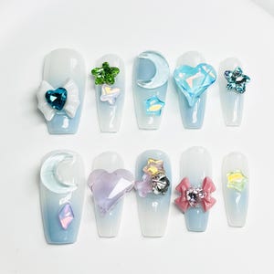 Op de afbeelding: Een set van tien kunstnagels, elk beschilderd met een verloop van lichtblauw naar wit. De nagels zijn versierd met diverse versieringen, waaronder strikken, harten, sterren en halve manen, allemaal in pastelkleuren en glinsterende strass-steentjes.