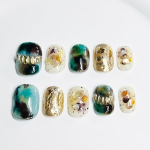Puede incluir: Conjunto de diez uñas postizas con una variedad de diseños. Las uñas presentan una mezcla de colores verde, marrón, dorado y blanco. Algunas uñas tienen un acento dorado, mientras que otras tienen un diseño de conchas marinas y purpurina.