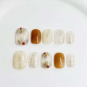 Puede incluir: Un conjunto de diez uñas artificiales con diversos diseños. Las uñas presentan una mezcla de colores claros, marrón y blanco, con elementos decorativos como purpurina plateada y patrones florales. Las uñas están dispuestas sobre una superficie blanca.