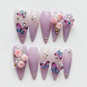 Puede incluir: Un juego de diez uñas postizas en forma de estilete con un degradado de purpurina lavanda y blanco. Las uñas están decoradas con rosas tridimensionales, mariposas iridiscentes, cuentas de perlas blancas y pequeños diamantes de imitación.