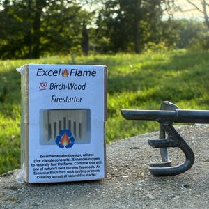 May include: A rectangular box of Excel Flame 100 Birch-Wood Firestarter with a white label featuring text and a flame logo, positioned next to a dark metal fire poker tool. The packaging includes a cutout window showing the wood contents inside.