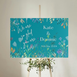 Może przedstawiać: Turkusowy niebieski znak powitalny na ślub z białymi wzorami kwiatowymi i tekstem "We are so glad you are here!"  Kate & Dominic 14th May 2024.
