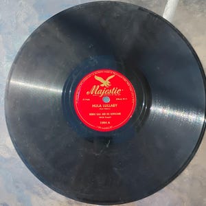 Puede incluir: Un disco de vinilo negro vintage con una etiqueta roja y blanca. La etiqueta dice "Majestic" con un emblema de águila y el título "Hula Lullaby" de Bernie Kaai and His Hawaiians. El disco está etiquetado 1064 A.