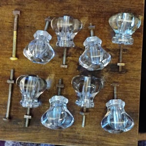 May include: A collection of clear glass cabinet knobs with metal screws and nuts. The knobs have a decorative, rounded shape and are arranged on a wooden surface. The hardware is vintage and suitable for furniture restoration.
