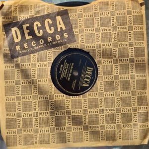 May include: Vintage Decca Records vinyl record in its original packaging. The packaging features a repeating pattern of the Decca Records logo. The record label is black and gold, with the Decca logo and song information visible.