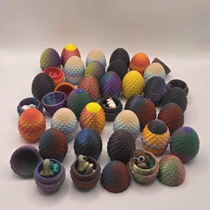 May include: A collection of decorative, colorful dragon egg-shaped containers. Some eggs are open, revealing small figurines inside. The eggs feature a variety of colors, including blue, green, orange, and purple. The containers are designed with a textured, scaled pattern.