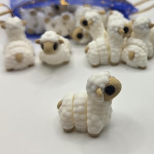May include: A collection of small, white sheep figurines with tan accents. The sheep have a textured, wool-like appearance and are arranged on a white surface. Some are in a blue mesh bag with gold string.