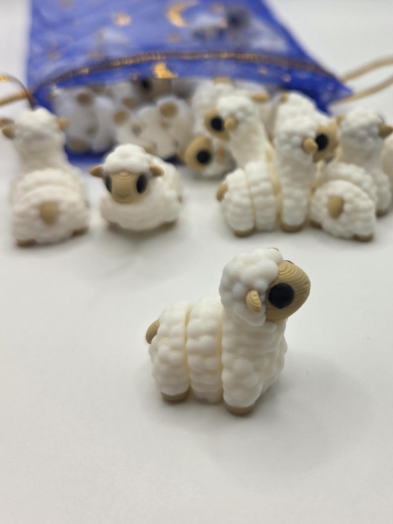 May include: A collection of small, white sheep figurines with tan accents. The sheep have a textured, wool-like appearance and are arranged on a white surface. Some are in a blue mesh bag with gold string.