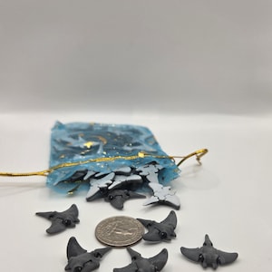 May include: A collection of small, gray stingray figurines, some with white accents, are scattered around a blue mesh bag with a gold drawstring. A US quarter coin is next to the figurines. The stingrays have a detailed design.