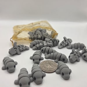 May include: A collection of small, gray manatee figurines scattered on a white surface, with a quarter for size comparison. Some figurines are in a small, gold mesh bag. The manatees have detailed features and black eyes.