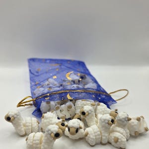 May include: A collection of small, white sheep figurines with tan accents, spilling from a blue mesh bag with gold star and moon details. The sheep are arranged on a white surface, creating a whimsical scene.