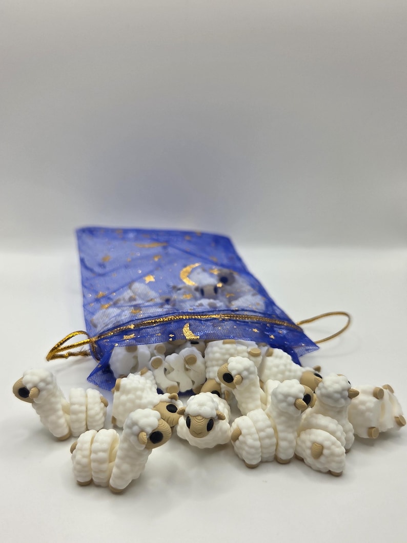 May include: A collection of small, white sheep figurines with tan accents, spilling from a blue mesh bag with gold star and moon details. The sheep are arranged on a white surface, creating a whimsical scene.