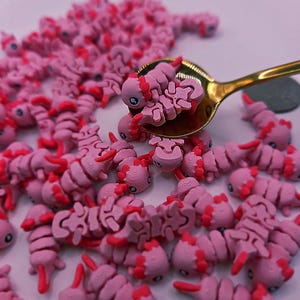 May include: A collection of pink axolotl-shaped charms with red accents, some held in a gold spoon. The charms are scattered across a white surface. The axolotls have detailed features.