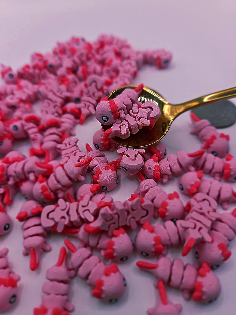 May include: A collection of pink axolotl-shaped charms with red accents, some held in a gold spoon. The charms are scattered across a white surface. The axolotls have detailed features.