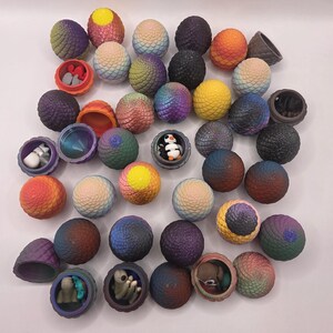 May include: A collection of colorful, textured, spherical objects, some opened to reveal small figurines. The spheres are in shades of purple, blue, orange, and yellow. Some spheres have a dragon egg design. The figurines include a penguin and small creatures.