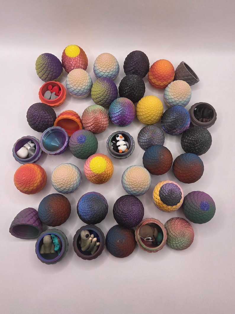 May include: A collection of colorful, textured, spherical objects, some opened to reveal small figurines. The spheres are in shades of purple, blue, orange, and yellow. Some spheres have a dragon egg design. The figurines include a penguin and small creatures.