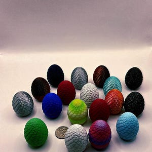 May include: A collection of decorative dragon eggs in various colors, including silver, blue, red, and green. Each egg has a textured, scaled surface. A US quarter is included for size reference.