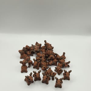 May include: A collection of small, brown, deer-shaped figurines. Each figurine has antlers and is detailed with carved lines. The figurines are scattered on a white surface, creating a playful and whimsical scene.