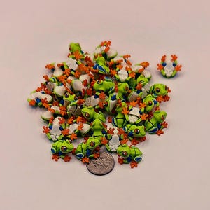 Peut inclure: Un ensemble de figurines de petites grenouilles vertes et blanches avec des pieds orange et des accents bleus. Les grenouilles sont &eacute;parpill&eacute;es sur un fond clair, avec une pi&egrave;ce de monnaie pour l'&eacute;chelle. Les grenouilles ont des yeux rouges.