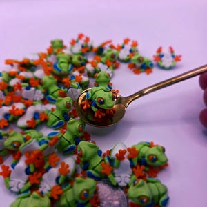 Peut inclure: Plusieurs figurines de petites grenouilles vertes avec des pieds orange et des d&eacute;tails bleus. Certaines grenouilles sont sur une cuill&egrave;re dor&eacute;e. Les grenouilles mesurent environ 2,5 cm de long.