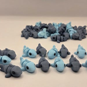 May include: A collection of small, stylized shark figurines in shades of blue and gray. The sharks are lying down, with a smooth, matte finish. Some are light blue, others are gray, and some are a mix of both colors. The figurines are grouped together.