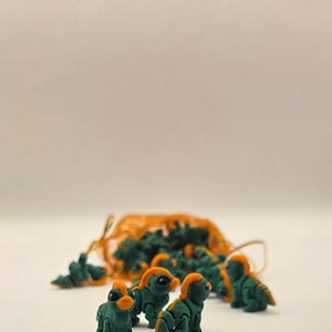 May include: A collection of small, green dinosaur figurines with orange accents. Several are arranged in front of an orange mesh bag filled with more figurines. The dinosaurs are on a white surface.