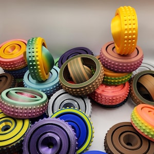 May include: A collection of colorful, 3D-printed fidget toys in various designs. The toys feature a circular shape with textured surfaces and come in a variety of colors, including yellow, pink, blue, and green.