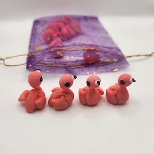 May include: Four small, pink flamingo figurines with black eyes and beaks are arranged in a row. A purple mesh bag with gold accents and additional flamingo figures is in the background.