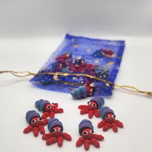 May include: A collection of handmade clay octopus figurines. Each octopus is red with a blue and purple hat, and white eyes. A blue mesh bag with gold string and colorful beads is in the background. The octopus figurines are arranged in a circle.