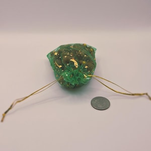 May include: A small green mesh bag filled with small, round, dark green objects and gold accents. The bag is tied with gold string and sits next to a coin on a white surface.