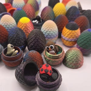 May include: A collection of colorful, dragon egg-shaped containers, some open to reveal small figurines like an owl and a ladybug. The eggs vary in color, including shades of blue, green, yellow, and orange, with textured surfaces.