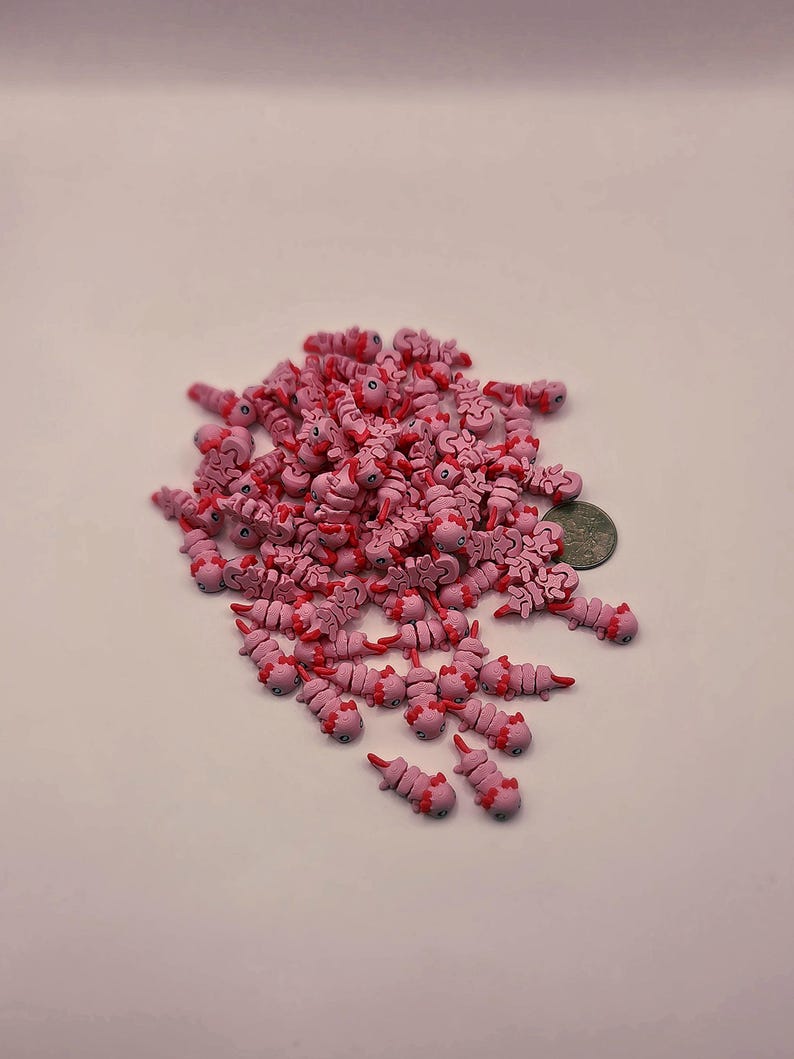 May include: A pile of small, pink axolotl-shaped charms with red accents. The charms are scattered on a white surface, with a coin visible for scale. The axolotls have detailed features, including tiny legs and gills.