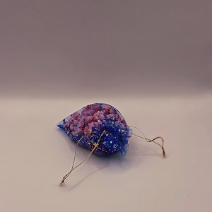 May include: A small, blue mesh bag filled with pink and purple decorative items. The bag has a drawstring closure and gold-colored accents. The contents appear to be small, possibly beads or decorative elements.