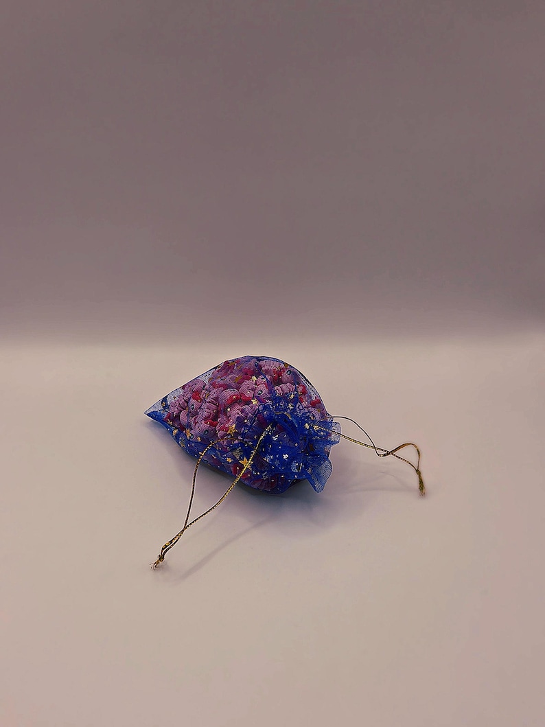 May include: A small, blue mesh bag filled with pink and purple decorative items. The bag has a drawstring closure and gold-colored accents. The contents appear to be small, possibly beads or decorative elements.