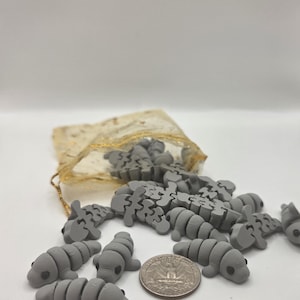 May include: A collection of small, gray manatee-shaped puzzle pieces scattered on a white surface, with a quarter coin for size comparison. A small, gold-colored drawstring bag is partially visible in the background.