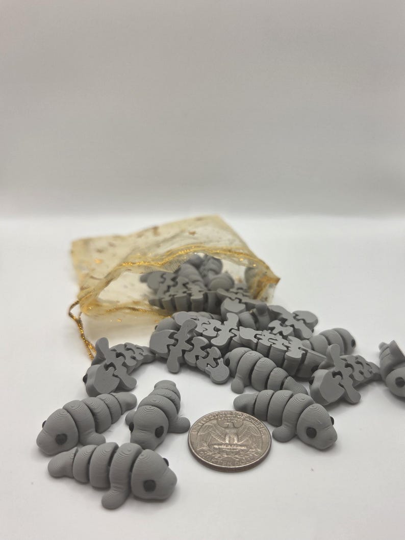 May include: A collection of small, gray manatee-shaped puzzle pieces scattered on a white surface, with a quarter coin for size comparison. A small, gold-colored drawstring bag is partially visible in the background.