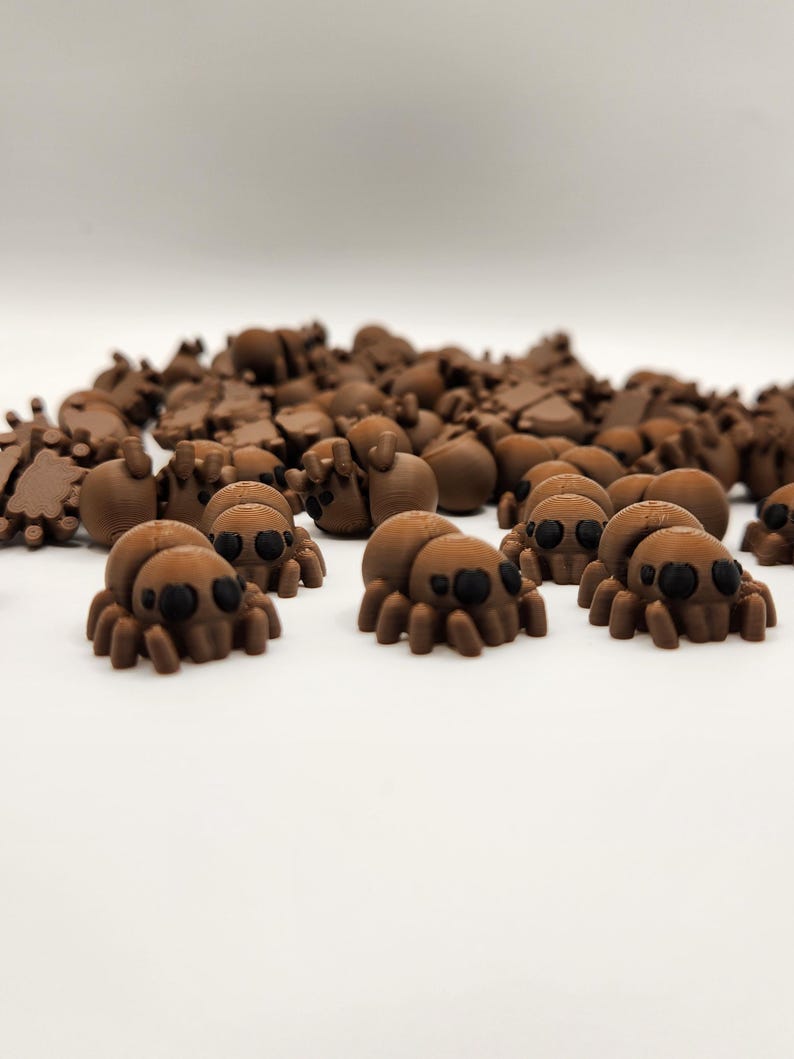 May include: A group of small, brown, 3D printed spiders with black eyes. The spiders are scattered on a white surface.