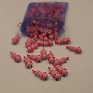 May include: Pink axolotl charms with red gills and small black eyes are scattered on a white surface. A blue mesh bag is in the background, filled with more of the same charms. These small, detailed axolotl figurines are likely for crafting or jewelry making.