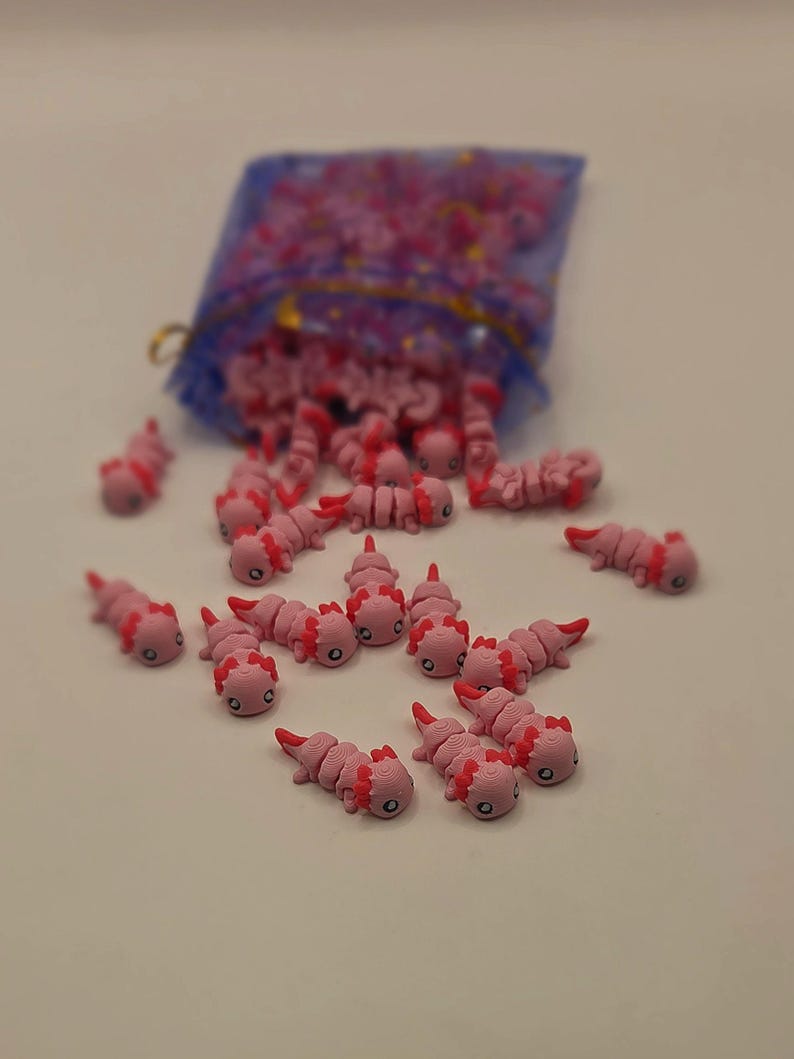 May include: Pink axolotl charms with red gills and small black eyes are scattered on a white surface. A blue mesh bag is in the background, filled with more of the same charms. These small, detailed axolotl figurines are likely for crafting or jewelry making.
