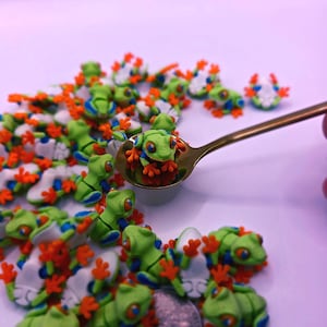 Peut inclure: Une collection de petites figurines de grenouilles vertes avec des pieds oranges et des d&eacute;tails bleus. Certaines grenouilles sont blanches avec des d&eacute;tails orange et bleu. Une cuill&egrave;re dor&eacute;e contient une grenouille. Les grenouilles sont &eacute;parpill&eacute;es sur une surface violet clair.