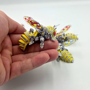 May include: A mechanical wasp figurine featuring a yellow and silver segmented body with metallic accents. The wings are translucent with red and yellow patterns, and the head has large red eyes. Additional similar wasp figurines are visible nearby.