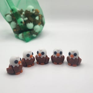 May include: Five small brown and white eagle figurines with black eyes and orange beaks. The figurines are arranged in a line on a white surface.