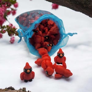Pu&ograve; includere: Un sacchetto di rete blu pieno di piccole figurine di cardinali rossi. Diversi cardinali si riversano dalla borsa su una superficie bianca.