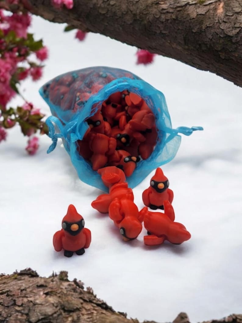 Pu&ograve; includere: Un sacchetto di rete blu pieno di piccole figurine di cardinali rossi. Diversi cardinali si riversano dalla borsa su una superficie bianca.