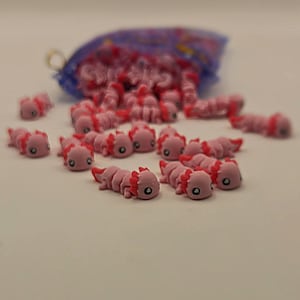 May include: A collection of small, pink axolotl figurines scattered on a white surface, with a blue mesh bag in the background. The axolotls have detailed features, including red gills and small black eyes. The figurines are likely made of plastic or resin.