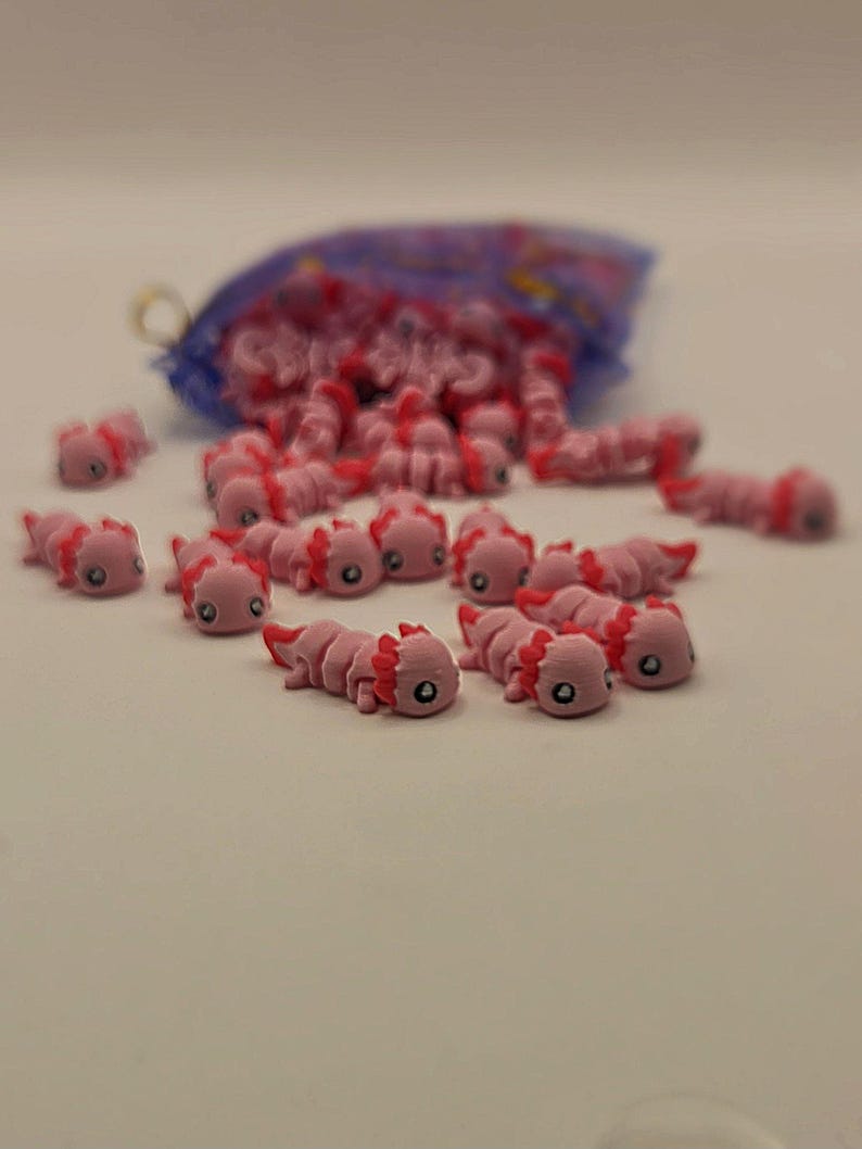 May include: A collection of small, pink axolotl figurines scattered on a white surface, with a blue mesh bag in the background. The axolotls have detailed features, including red gills and small black eyes. The figurines are likely made of plastic or resin.